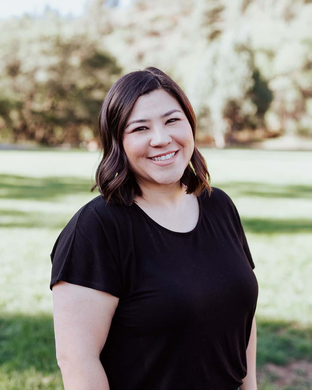 Smiling woman with dark hair in a black shirt standing in a sunlit park with trees in the background