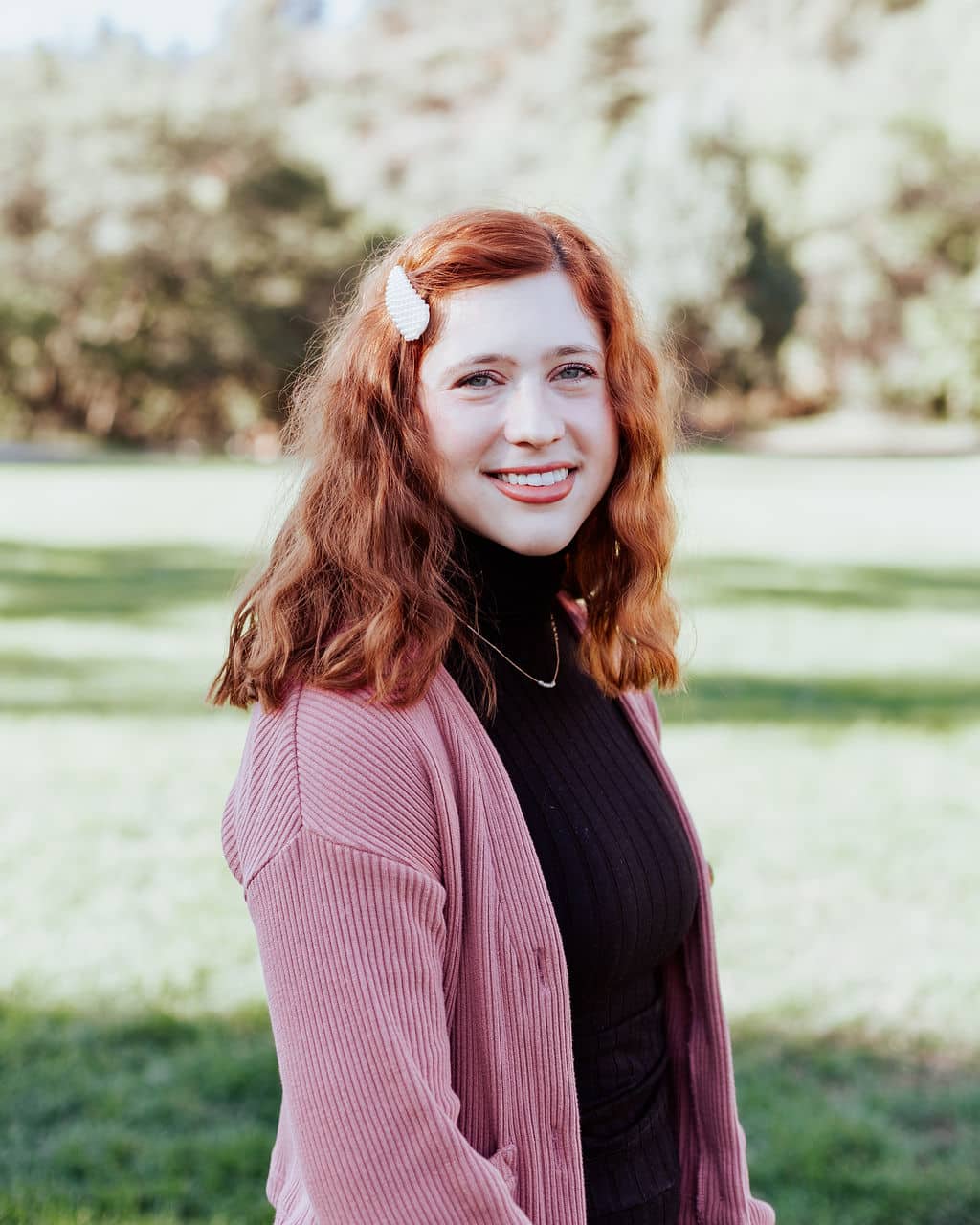 Young woman with red hair smiling at the camera in a park, wearing a pink cardigan over a black ribbed top.