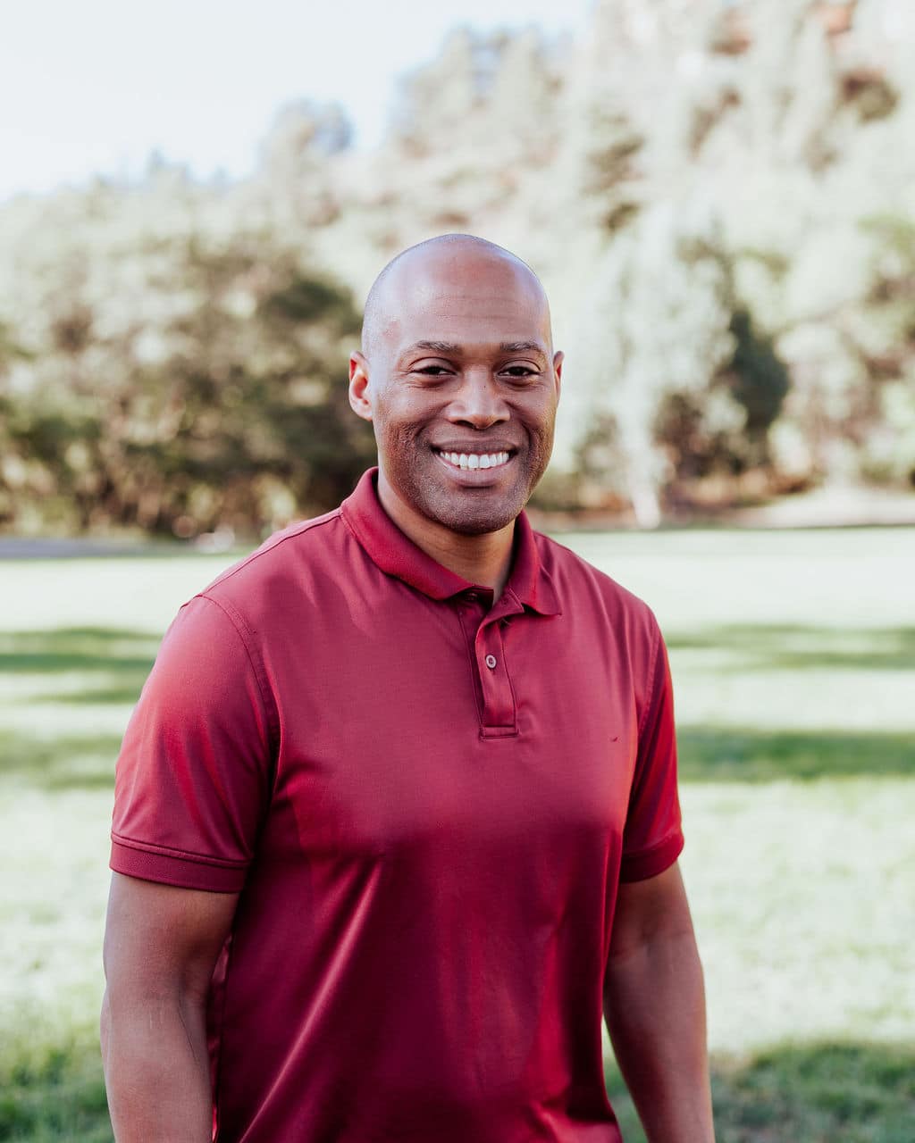 Smiling man in a maroon polo stands in a sunny park with green grass and trees in the background in a portrait.